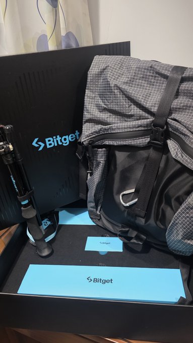 An open Bitget-branded box containing a black and gray checkered backpack, a tripod, and Bitget-branded cards and packaging materials.