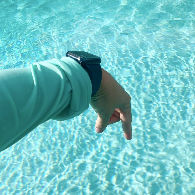 A person's arm wearing a black smartwatch and light-colored sleeve extends over a bright blue swimming pool, with their hand making a small gesture.
