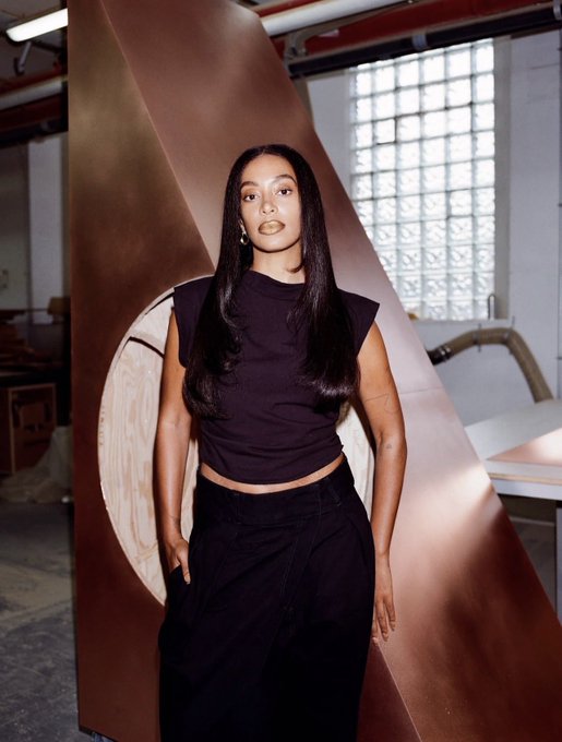 Solange poses in a sleek black outfit featuring a structured shoulder top and draped skirt, standing before a large copper-toned sculptural installation in an industrial loft setting with glass block windows.