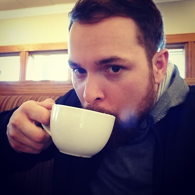 A close-up selfie shows a bearded man drinking from a large white ceramic mug, with windows and a wooden booth visible in the background.