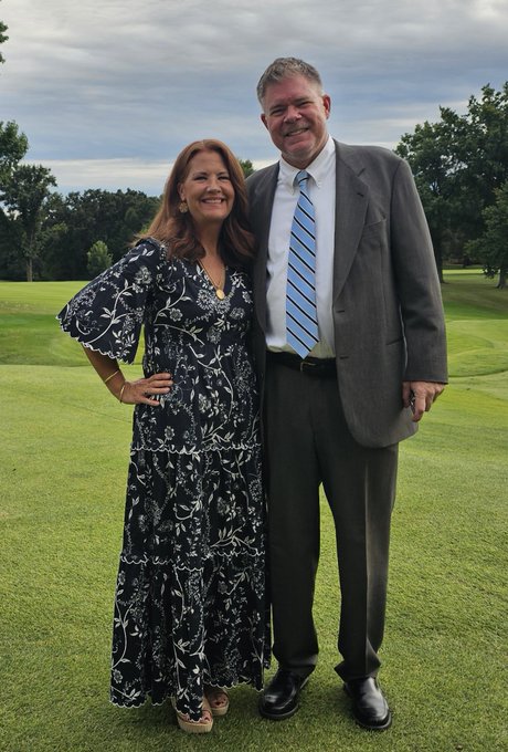 A smiling middle-aged couple poses together on a green lawn, with the woman wearing a floral navy dress and the man in a gray suit with a striped tie.