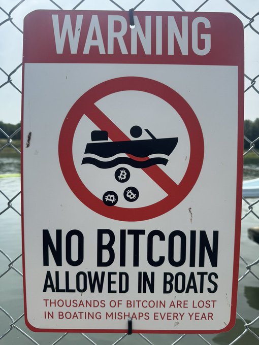 A humorous warning sign on a chain-link fence reads "WARNING: NO BITCOIN ALLOWED IN BOATS - THOUSANDS OF BITCOIN ARE LOST IN BOATING MISHAPS EVERY YEAR," featuring a crossed-out boat dropping Bitcoin, Bitcoin Cash, and Dogecoin tokens into the water. The satirical sign pokes fun at crypto holders who lose their hardware wallets (and thus their coins) to lakes and oceans, highlighting the absurd yet relatable phenomenon that the post is critiquing.