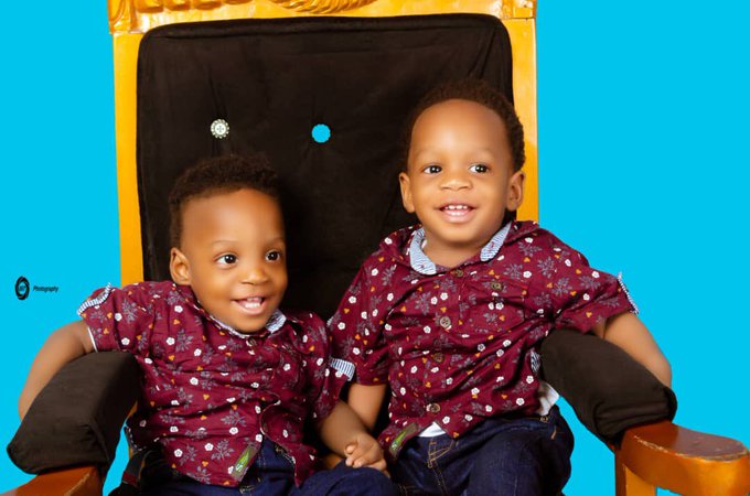 A professional portrait of two smiling young boys with short hair, wearing matching maroon patterned shirts and jeans, posed together in front of a yellow and black backdrop.