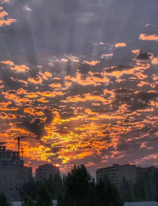 A dramatic sunset sky with orange and grey clouds over a city skyline featuring construction cranes and apartment buildings.