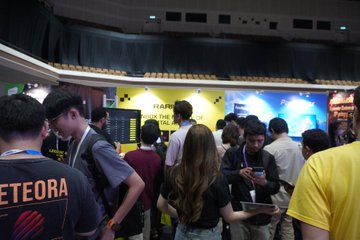 A crowd of attendees gathers at a busy booth featuring yellow "Happy" signage, with one person wearing a "ETEORA" jersey in the foreground, capturing the overwhelming turnout at the event.