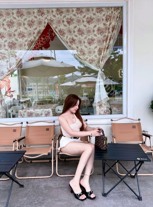 A woman in a short white dress sits at a cafe table outside a window decorated with floral curtains, holding a Louis Vuitton bag.
