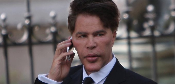 A man in a dark suit speaks on a cell phone with a concerned expression, standing in front of an ornate iron gate. The image doesn't directly relate to the post about the crypto trading platform and its funding from Ribbit Capital, though the phone call imagery could symbolize business negotiations or announcements.