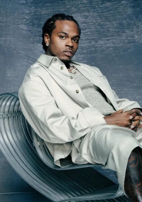 A man with a serious expression sits in a modern silver chair against a textured blue backdrop, wearing a cream-colored leather jacket, gray sweater, and white pants, with visible tattoos on his leg. The accompanying post lists various artists and albums, and the image likely represents "Johnny Venus (of EARTHGANG)" promoting his "THE 11TH HOUR" project from the featured list.
