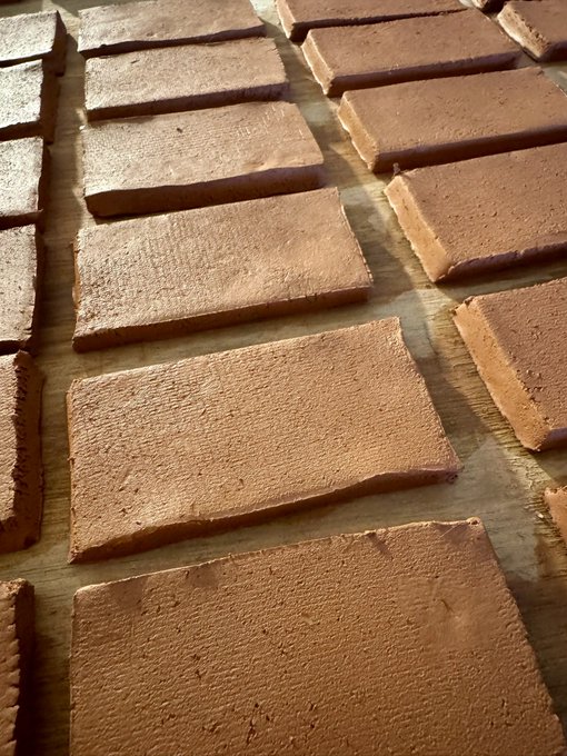 The image features a large number of rectangular bricks made from clay, arranged neatly across a wooden surface. The bricks are of varying sizes and shapes, creating an interesting visual effect. The background appears to be a simple gray color, which contrasts with the warm tones of the bricks. The text in the image reads "GM, Tezos fam! Great news — this month we’re launching 12 collections in partnership with Maskay Gallery on @objktcom". This suggests that there is an art exhibition or event related to clay and brickwork taking place at a gallery named "@maskaygallery" and it has been posted on social media.