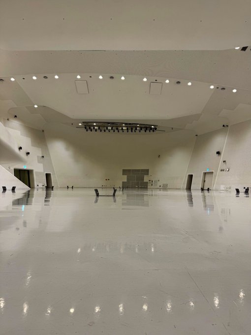 An empty, modern performance hall with a glossy white floor, curved walls, and overhead stage lighting — the perfect blank canvas setting where the "Breakpoint" event will take place.
