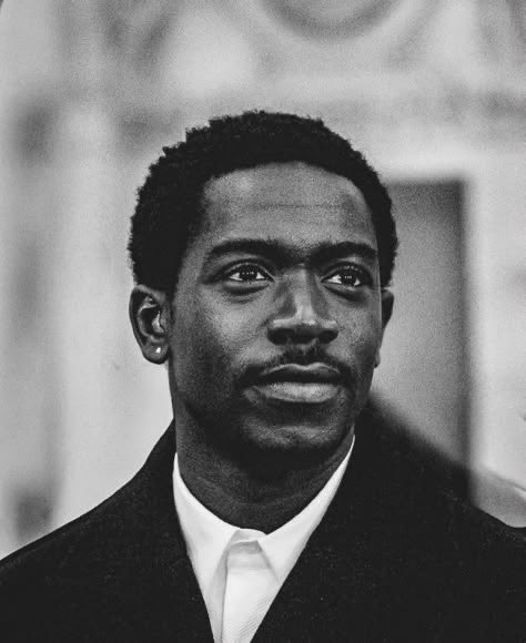 A black and white portrait photograph shows a young man with short dark hair, wearing a suit jacket and white collared shirt, gazing upward.