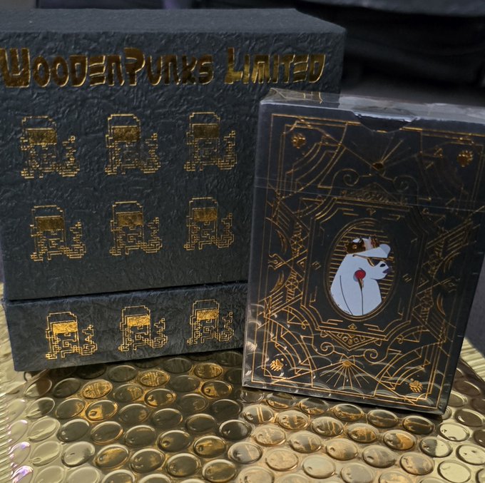 A deck of 'WoodenPunks Limited' playing cards with pixel art and ornate gold designs sits next to its black box, displayed on a gold hammered tray.