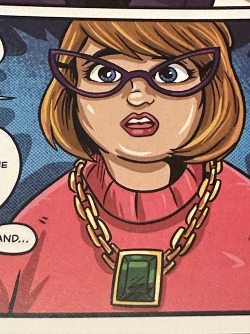 Comic book panel showing a woman with a brown bob haircut, purple cat-eye glasses, and a large gold chain necklace featuring a green gemstone pendant, wearing a pink top with a partially visible speech bubble ending in "...AND..."
