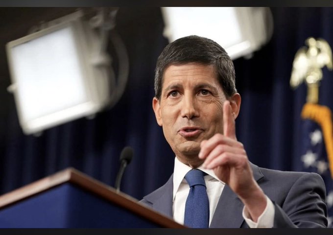 A man in a suit and blue tie speaking at a podium, gesturing with his finger raised, with a dark blue curtain and an American flag in the background.