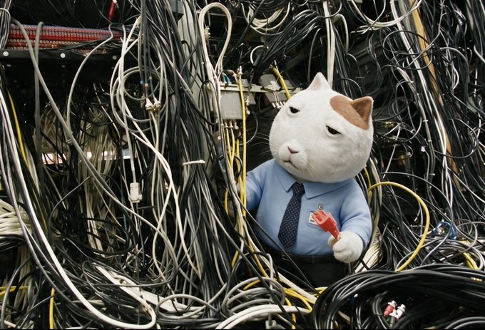 A plush cat wearing a blue shirt and tie holds a red connector while surrounded by a massive tangle of cables.