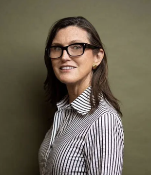 Cathie Wood, CEO of ARK Invest, wearing black-rimmed glasses and a striped blouse, shown in a professional portrait. Her image accompanies a bullish analysis on Bitcoin's recent breakout against gold, where she highlights the BTC-gold ratio reaching 18 and Bitcoin's 22% monthly outperformance over gold's 2% gain.
