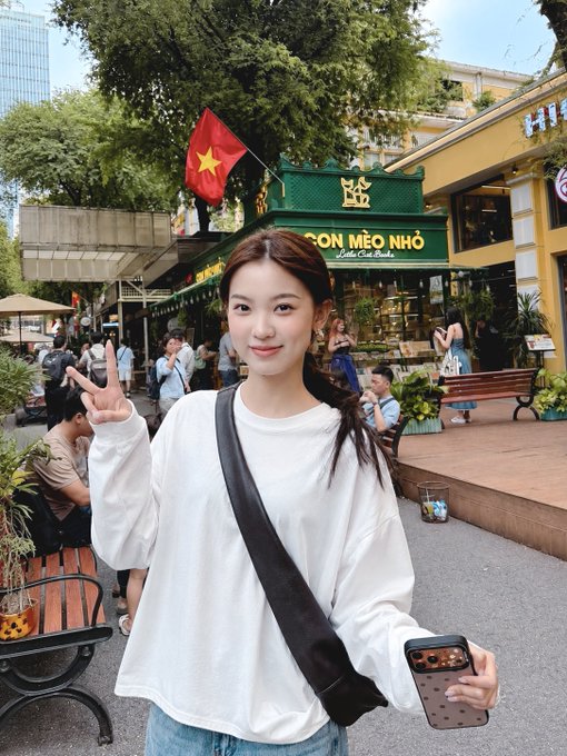 A young woman with long brown hair, wearing a white long-sleeved shirt and a black crossbody bag, smiles and flashes a peace sign while holding a smartphone in front of a yellow cafe named 'CON MÈO NHỎ - Coffee - Cake - Brunch' with a Vietnamese flag flying nearby.