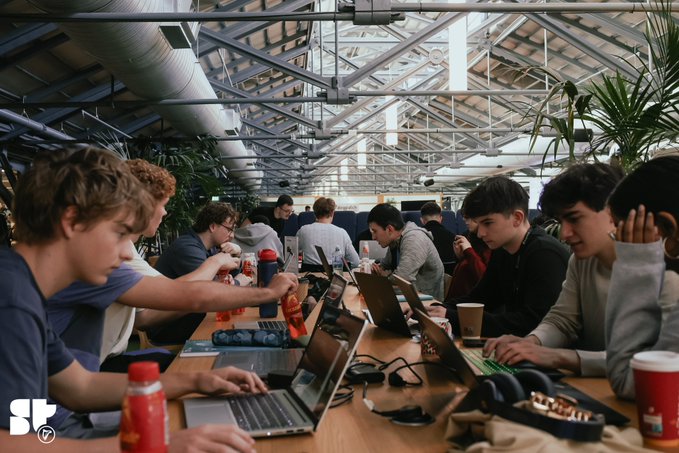 A group of young people working on laptops at a long table in a greenhouse-style coworking space with a glass roof and exposed ductwork.