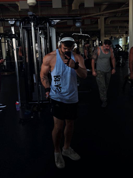 The image shows a man wearing headphones, standing in front of a weight machine at a gym. He appears to be listening to music or an audio program while working out. Another person can be seen walking by him, possibly going about their own workout routine. The scene suggests that this is a typical day at the gym for people who are exercising and using various equipment like treadmills, dumbbells, and weight machines.