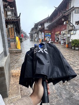 A hand holds a closed black umbrella with 'XO' text on a rainy traditional street with stone pavement and old buildings.