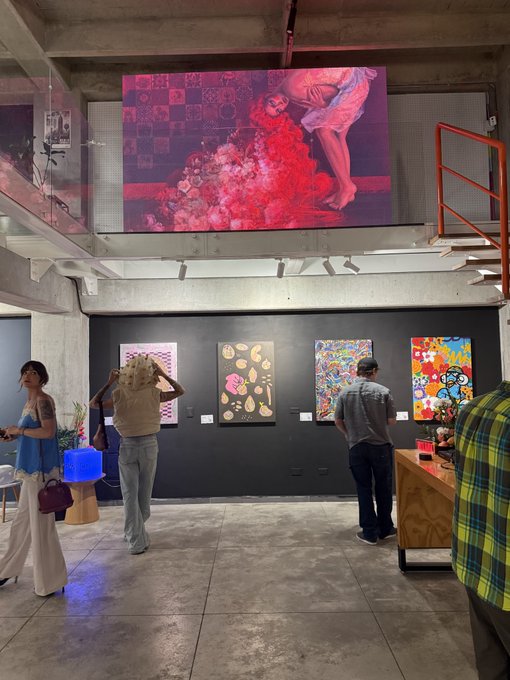 An art gallery interior with visitors viewing colorful artworks displayed on a black wall, while a large illuminated screen shows a red-toned figure with flowers hanging from the ceiling.