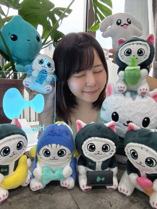 A young woman with dark hair smiling with her eyes closed, surrounded by a collection of cute plush toys including a blue water droplet and several cats in hoodies, with a Windows logo sign visible.