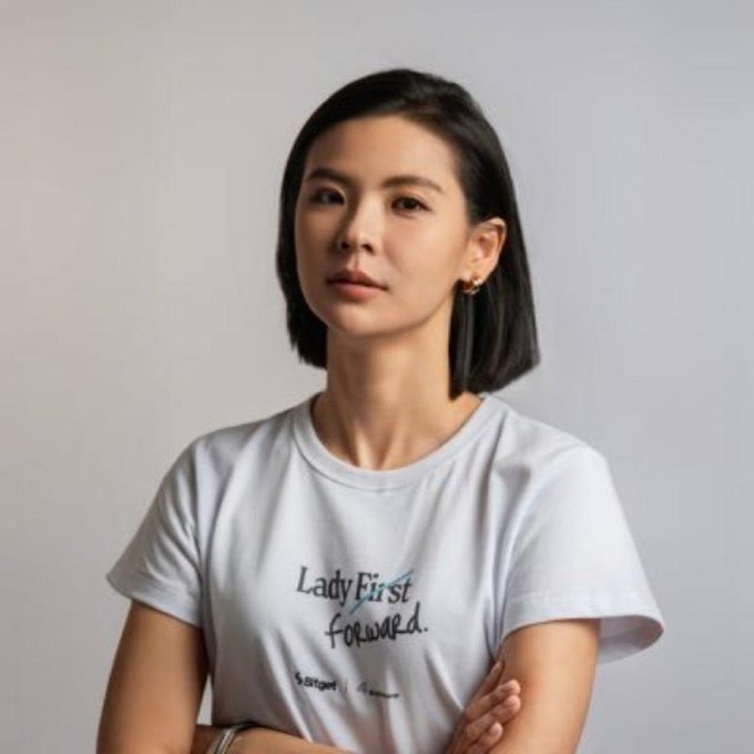 A woman with a short black bob haircut wears a white t-shirt that reads 'Lady First forward.' with arms crossed.