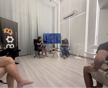 A small room shows people watching a Bitcoin chart on a screen, with a 'BTC' Bitcoin logo banner in the corner.