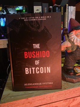 A book titled 'The Bushido of Bitcoin' by Aleksandar Svetski, with a foreword by Michael Saylor, sits on a shelf next to a small gnome figurine.