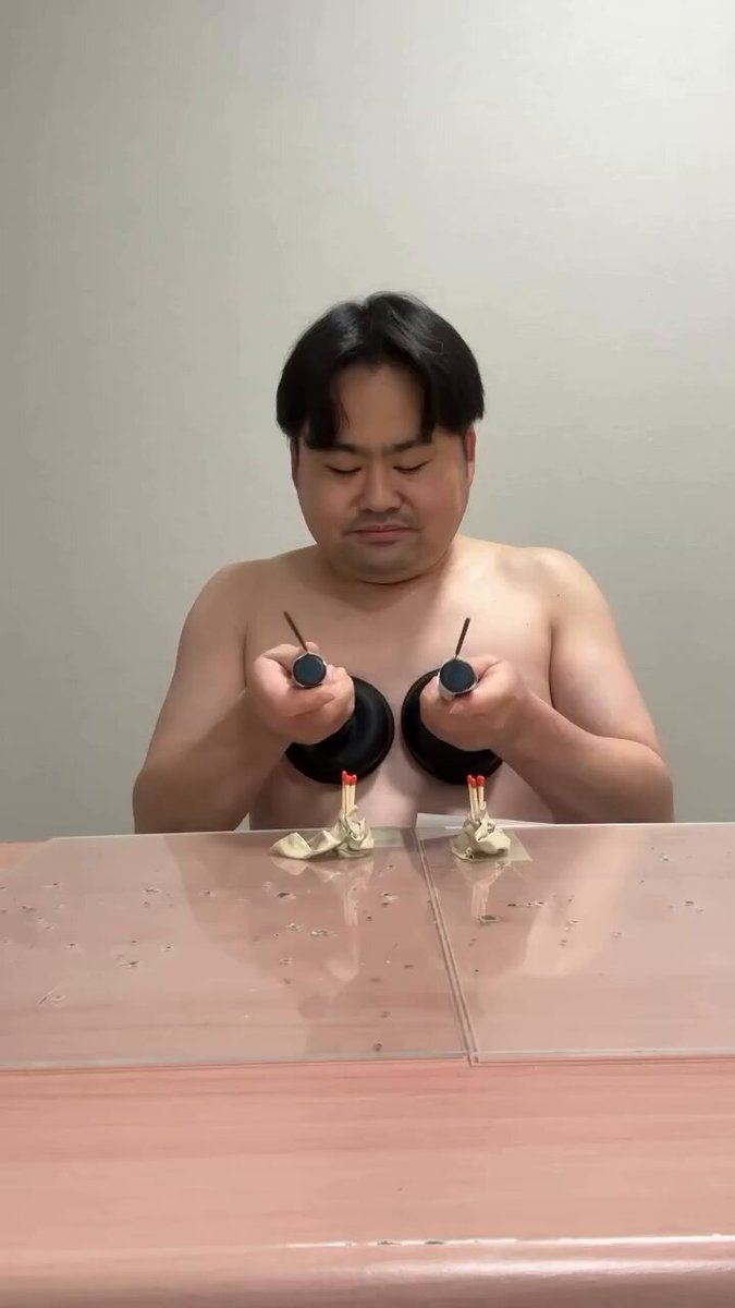 The image features a shirtless man sitting at a table with two candles burning on either side of him, creating an unusual and intriguing scene. The background appears to be a plain white wall, which contrasts with the candles and the man's attire. The caption reads "My friend from japan @ Liberal_Shuwski promise will do this when $MON back to ath", suggesting that the image is related to a social media post or a personal message shared by the man in question.