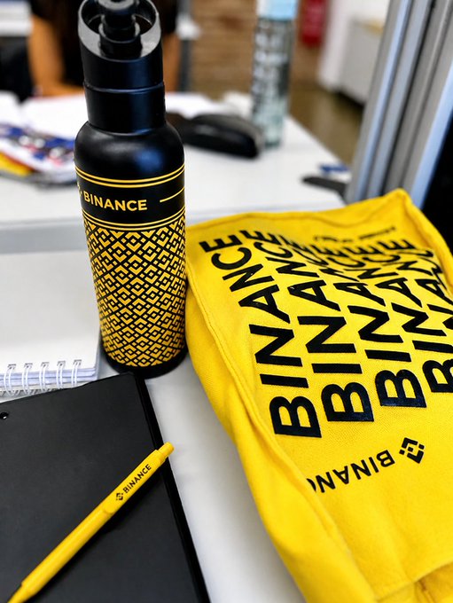 A black and yellow Binance branded water bottle and a yellow shirt printed with repeating 'BINANCE' text sit on a table next to a yellow Binance pen.