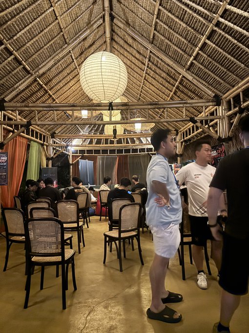 A group of people gather inside a rustic building with a bamboo/palm thatched roof, paper lanterns, and rows of chairs, with a man in a light blue shirt and white shorts standing in the foreground.
