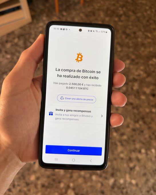 The image shows a person holding a smartphone displaying an app that allows them to invest in Bitcoin. The phone's screen displays information about the Bitcoin transaction, including the amount invested (2,500€) and the current value of the Bitcoin being invested (0.41).