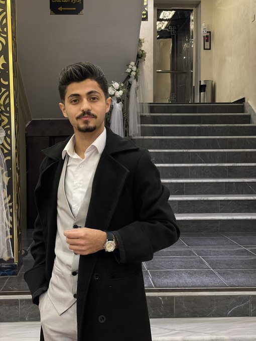 A young man in a black coat and white shirt stands on a marble staircase, wearing a wristwatch and looking at the camera in what appears to be an elegant indoor venue.