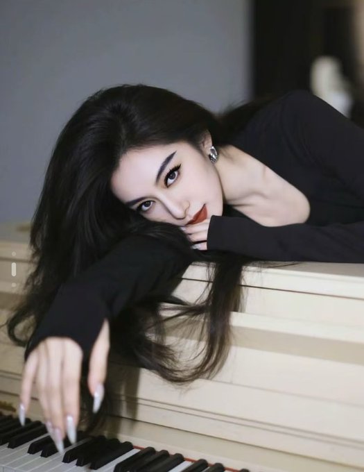 An elegant portrait of a woman with long dark hair, wearing a black top and long white nails, resting on a piano keyboard.