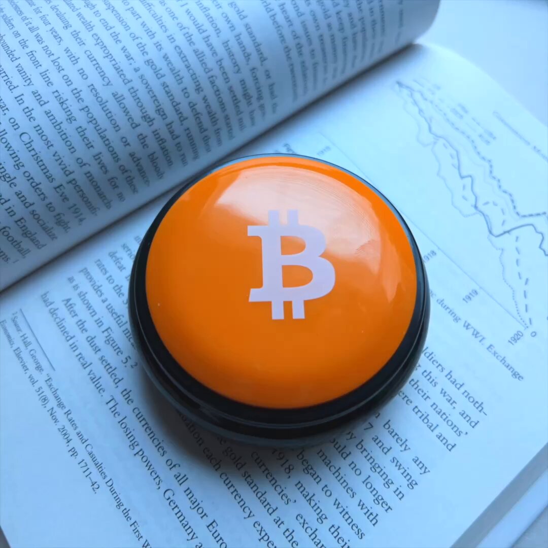 The image features a small orange button with a white Bitcoin symbol on it placed on top of an open book. The book appears to be a page from a magazine or newspaper, possibly a financial one, as indicated by the presence of graphs and charts in the background. The button is positioned near the bottom right corner of the image.