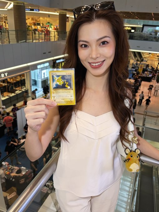 A smiling woman in a white top stands in a shopping mall holding a Pikachu trading card, with sunglasses on her head and a yellow character keychain on her bag.