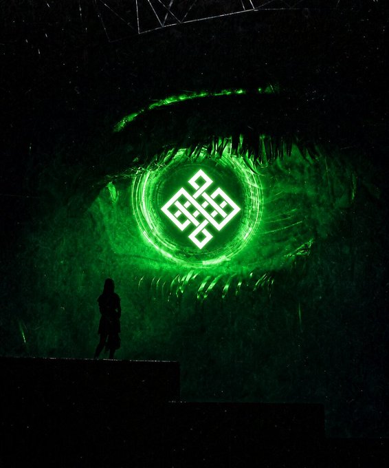 A dramatic stage scene features a large green illuminated eye design with a geometric symbol in the center, while a silhouetted figure stands below.