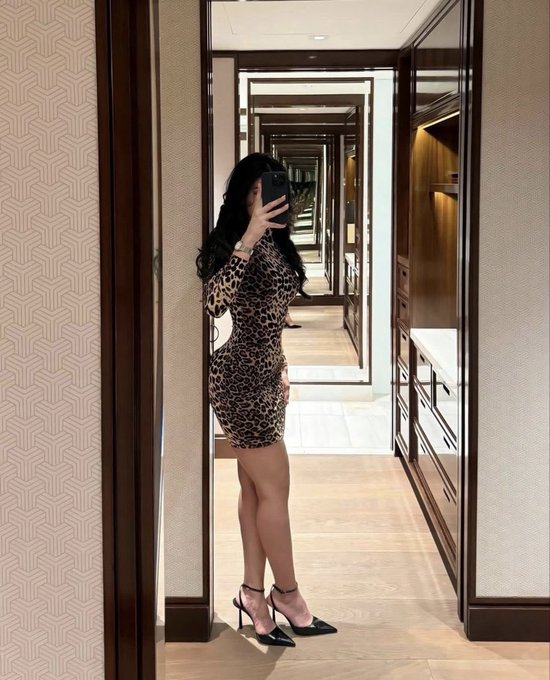 A mirror selfie of a woman wearing a leopard-print bodycon dress and black strappy heels, standing in an elegant hallway.