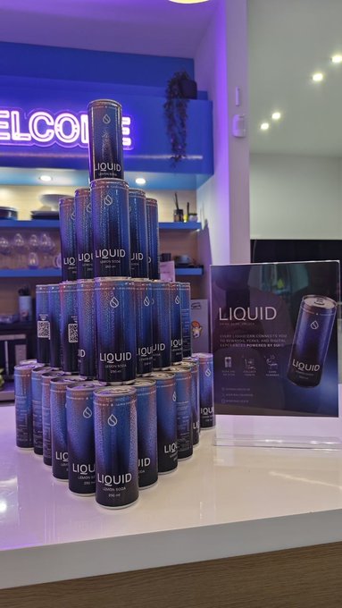 A pyramid display of "LIQUID" sparkling water cans sits on a counter alongside a promotional sign, tying in with the post about blockchain-enabled sparkling water featured at the SuiHub DeFi event hosted by @SuiCommunity.