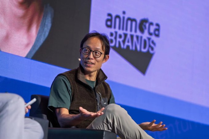 A man with glasses wearing a green shirt and dark fleece vest speaks on a stage, with 'animoca BRANDS' displayed on the blue screen behind him.
