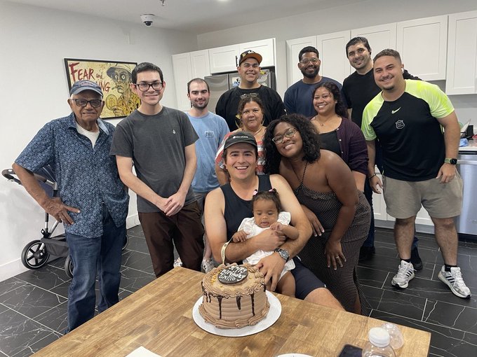 A large multi-generational family of around ten people poses for a photo around a wooden table with a chocolate birthday cake, in a kitchen setting with white cabinets.