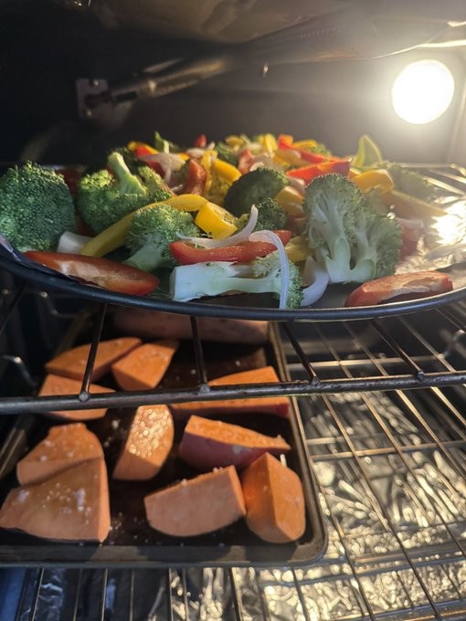 A view inside an oven showing a tray of chopped vegetables (broccoli, bell peppers, onions) on the top rack and a tray of sweet potato wedges cooking on the rack below.