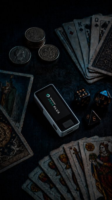 A product photograph showing a Tangem Wallet Plus hardware crypto wallet displayed on a dark surface alongside vintage tarot cards, coins, and dice in moody lighting.