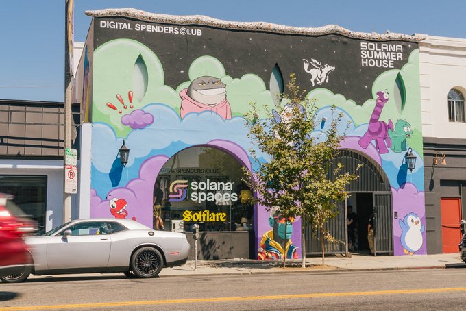 A colorful mural on a storefront featuring whimsical cartoon characters—including a shark in a hoodie, a penguin, and a pig—adorns the "Solana Spaces" building, with signage reading "Digital Spenders Club," "Solana Summer House," and "Solflare." The vibrant street art brings a playful, crypto-themed cultural vibe to the neighborhood, blending digital finance branding with creative expression.