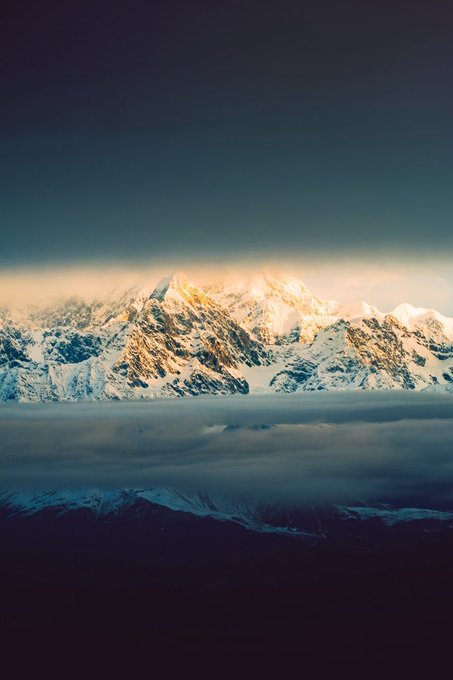 A scenic photograph of snow-capped mountains rising above a thick layer of clouds, illuminated by golden sunset light against a dark blue sky.