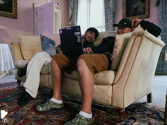 Two men relax on a cream-colored couch in an ornate room, one using a laptop while the other sits beside him wearing a cap and camouflage Crocs.