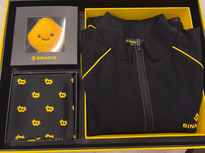 An opened Binance gift box containing a black and yellow jacket with Binance branding, a small orange Binance mascot plushie in a display frame, and a pair of black socks with yellow Bitcoin logos.