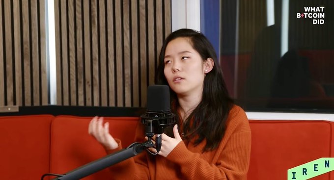 A woman named Iren speaks into a microphone during a 'What Bitcoin Did' podcast episode, seated on a red couch.