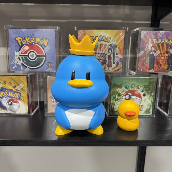 A blue penguin-like Pokémon figure wearing a yellow crown sits on a shelf next to a yellow rubber duck, with Pokémon trading card packs displayed in the background.